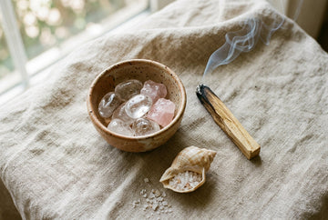Oczyszczone kamienie — kryształ górski i kwarc różowy w ceramicznej misce obok dymiącego palo santo i muszli z solą morską